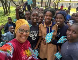 Akiliah Perez smiles for a selfie with a group of Kenyan schoolgirls holding small blue gift pouches, with more children gathered outdoors behind them.