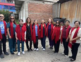 Ten people in scrubs and casual clothing stand in a row in a paved courtyard, each wearing a red scarf, in front of a brick wall and a corrugated metal fence. One man wears a Loma Linda University Mission Service t-shirt.