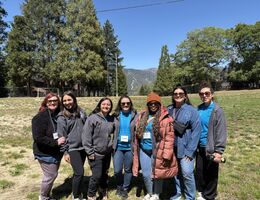 A group of seven Camp Good Grief counselors smile together on an open field surrounded by tall pine trees and mountain views.