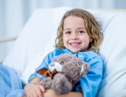 A child leans against a raised hospital bed, holding a teddy bear