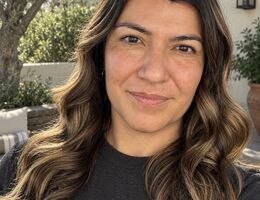 Close-up headshot of Dr. Danielle Lascano with long wavy brown hair smiling softly outdoors, with greenery and a stone building in the background.