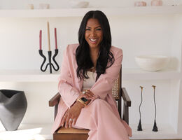Dr. Allycin Powell-Hicks smiling in a blush pink suit, seated in a cane-back armchair in a styled, modern interior.