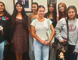 Group of nine female students smiling together in a wood-paneled office, standing in front of framed portraits on the wall.