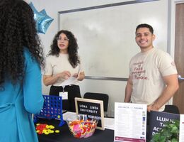 Two Loma Linda University Human Resources representatives speak with a student at their booth during the 2026 SBH Job Fair. The table displays a Connect Four game, candy, and informational materials including a 2025 Facts and Figures sheet.