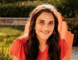 Dr. Lorraine Benuto, a woman with long dark wavy hair, smiling while seated in an orange Adirondack chair outdoors, wearing a red top and hoop earrings, with greenery in the background.