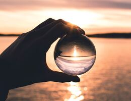 A hand cradles a glass orb reflecting a peaceful sunset over calm water, symbolizing clarity, reflection, and inner calm.