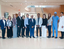 A group of 14 researchers and faculty in business attire, scrubs, and a white coat pose together in a bright hospital lobby with a spiral staircase in the background.