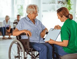 An older woman in a wheelchair speaks with a younger woman holding a clipboard
